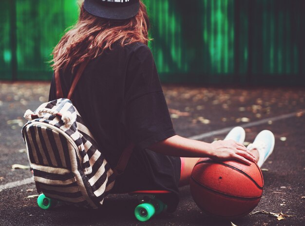 Closeup photo basketball ball with girl sitting on plastic orange penny shortboard on asphalt