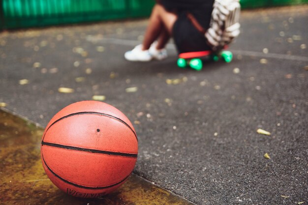 Closeup photo basketball ball with girl sitting on plastic orange penny shortboard on asphalt