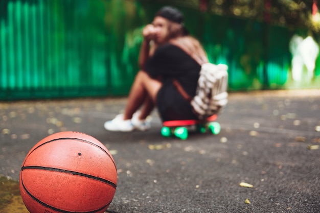 Closeup photo basketball ball with girl sitting on plastic orange penny shortboard on asphalt