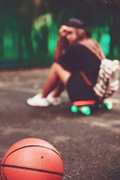 Closeup photo basketball ball with girl sitting on plastic orange penny shortboard on asphalt