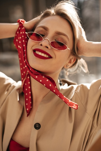 Free photo closeup portrait of attractive lady with gorgeous makeup smiling outside blonde woman in red sunglasses and beige trench coat touches hair