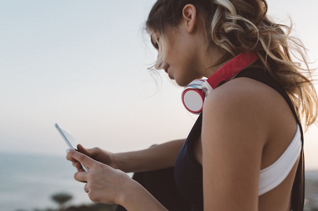 Free photo closeup portrait attractive sportswoman with headphones on neck, long curly hair chatting on phone, enjoying sunrise on seafront. cheerful mood, lovely summer time.