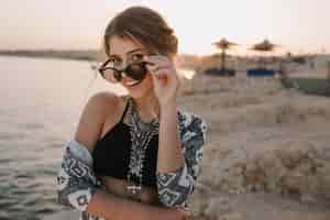 Free photo closeup portrait of attractive young girl taking off stylish sunglasses, on sunset, on beach with sensual look. wearing trendy black top, necklace, cardigan with ornaments. resort, vacation, holiday.