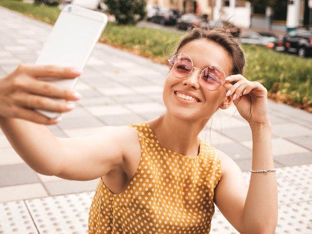 Closeup portrait of beautiful smiling brunette girl in summer hipster yellow dress. Model taking selfie on smartphone.Woman making photos in warm sunny day in the street in sunglasses