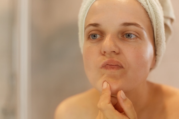 Closeup portrait of beautiful woman looking at her face, trying find acne, posing with naked shoulders and towel over head, doing morning beauty procedures.