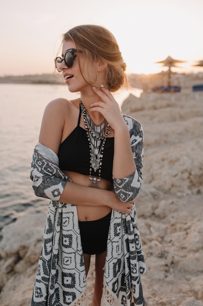 Free photo closeup portrait of beautiful young girl on beach, enjoying sunset, sensualy looking to side. wearing trendy black swimsuit, bikini, stylish sunglasses, necklace, cardigan, cape with ornaments.