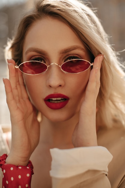 Free photo closeup portrait of blonde young woman with red lips putting on bright stylish colorful sunglasses elegant lady looks into camera outside