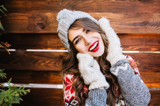 Free photo closeup portrait brunette beautiful girl with long hair in knitted grey hat and winter sweater on wooden . she touching face with hands in gloves and smiling .