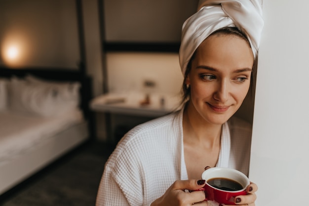 Free Photo closeup portrait of charming lady in white bathrobe and towel posing in bedroom with cup of morning coffee.