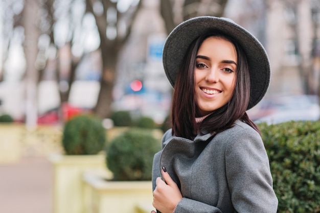 Free photo closeup portrait fashionable young  woman in grey hat, coat walking on street in city park. brunette hair, smiling, cheerful mood, elegant outlook.