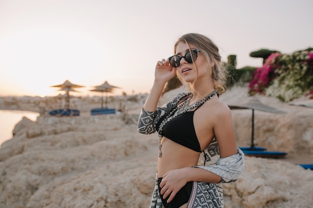 Free photo closeup portrait of gorgeous young girl with slim body wearing stylish black swimsuit, necklace, cardigan, cape with ornaments. beach, rocks, sunset.