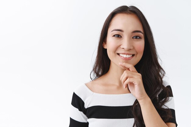 Closeup portrait happy tender young asian girl in striped tshirt touching chin and smiling toothy with delighted or satisfied expression gazing camera interested or intrigued white background