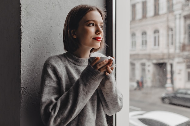 Closeup portrait of lady in gray wool sweater with red lipstick enjoying tea near window.