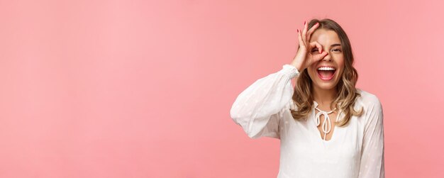 Closeup portrait of perfect goodlooking blond girl in white dress looking through okay sign and smil