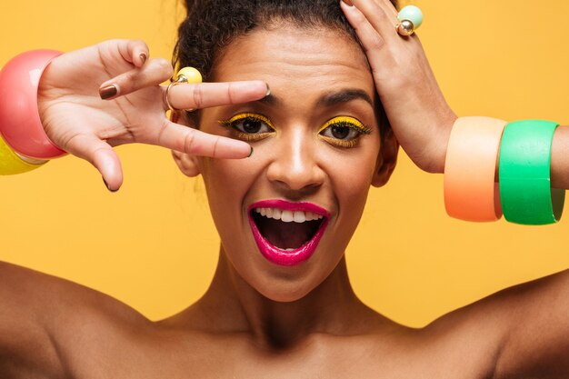 Closeup portrait of playful mulatto woman with yellow eyelids and pink lips gesturing two fingers at eye and looking on camera, isolated