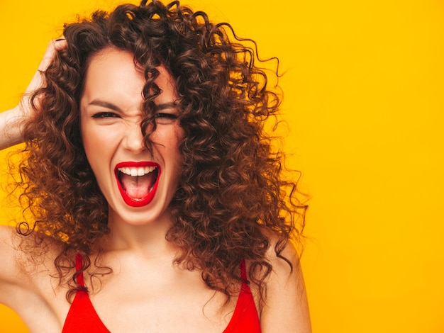 Free photo closeup portrait of sexy young beautiful smiling female carefree woman isolated on yellow wall in studio positive brunette model with afro curls hairstyle in sunglasses screams and shouts