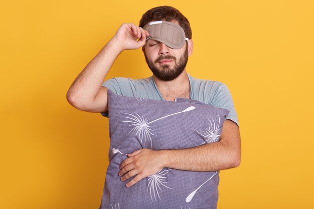 Closeup portrait of sleepy man with blindfold on eyes, holding pillow in hands, opens sleeping mask, keeping eyes closed