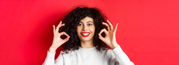 Free photo closeup portrait of smiling woman with curly hair and red lips showing okay gesture and looking sati