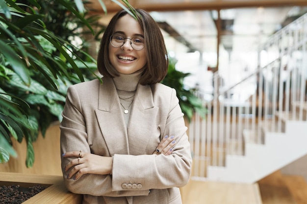 Closeup portrait of successful happy smiling young woman in beige jacket and glasses standing in lobby or office reception greeting business client with pleasant grin inviting to company