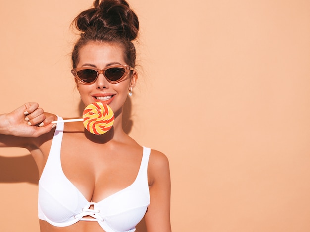 Free photo closeup portrait of young beautiful sexy smiling woman with ghoul hairstyle. trendy girl in casual summer white swimsuit in sunglasses.hot model isolated on beige.eating,biting candy lollipop