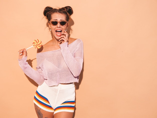 Free photo closeup portrait of young beautiful sexy smiling woman with ghoul hairstyle. trendy girl in casual summer white swimsuit in sunglasses.hot model isolated on beige.eating,biting candy lollipop