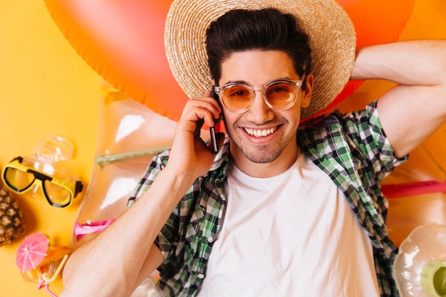 Closeup portrait of young guy in orange glasses and straw hat. Man in T-shirt is resting on inflatable mattress and talking on phone.