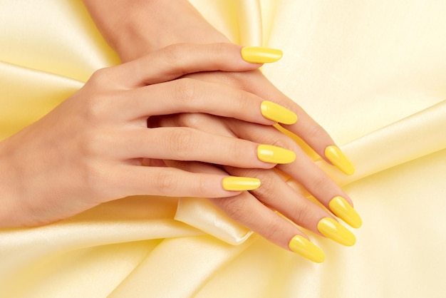 Closeup shot of a female's hands with yellow nail polish on a yellow silk fabric