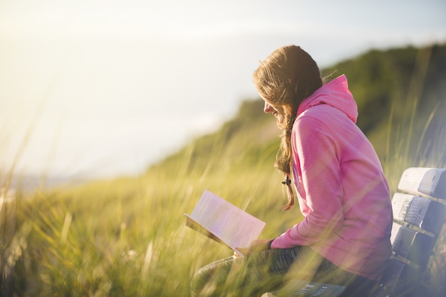 Closeup shot of a female sitting on the bench while reading the bible in a grass field