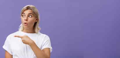 Free photo closeup shot of intrigued interested attractive adult blonde female in white tshirt folding lips staring and pointing left with curious and thrilled expression over purple background