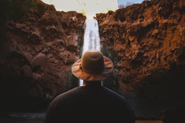 Free photo closeup shot of a person wearing brown hat facing a waterfall flowing down the hills