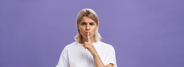 Free photo closeup shot of serious strict goodlooking caucasian female with blond hair shushing at camera displeased and disappointed frowning holding index finger over folded lips in shush sign