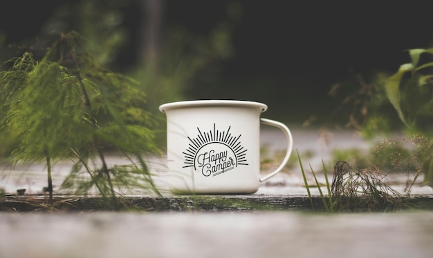 Closeup shot of a white metal cup with "Happy Camper" writing on a wooden surface near green plants