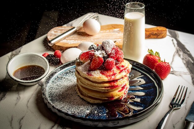 Closeup of a stack of appetizing pancakes topped with strawberries and ingredients in the background