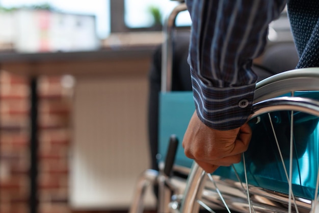 Free Photo closeup of startup employee hand holding rim of wheelchair wheel to move in front of desk with laptop with sales charts. focus on african american man living with disability moving around office.