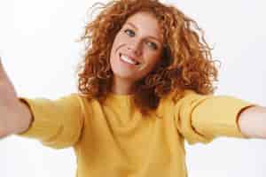 Free photo closeup tender modern cute redhead curly woman in yellow sweater enjoying warm sunny winter days taking picture of herself using smartphone camera make selfie pull hands forward