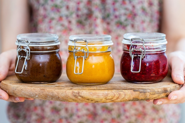 Closeup view of a woman holding a wooden plate with plum, apricot, raspberry jam glass jars