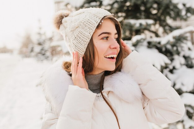 Free Photo closeup winter portrait young beautiful woman in white knitted hat expressing to side on street full with snow.