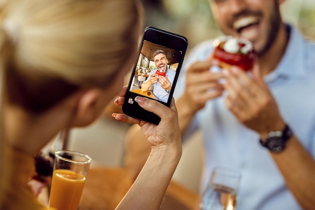 Closeup of woman taking picture of her boyfriend who is eating donut in a cafe