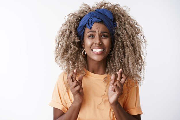 Closeup worried and anxious troubled young hopeless africanamerican curly blond woman in orange tshirt headband clench teeth desperate look camera cross fingers good luck praying