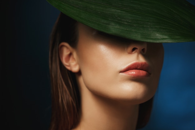 Closeup of young woman covering face behind fresh green leaf.