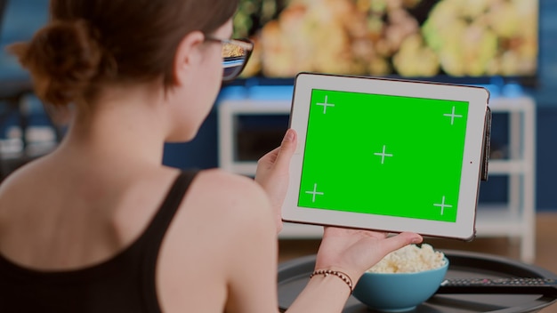 Closeup of young woman holding digital tablet with green screen watching webinar sitting on sofa in front of tv. Girl looking at touchscreen device with chroma key attending online course at home.