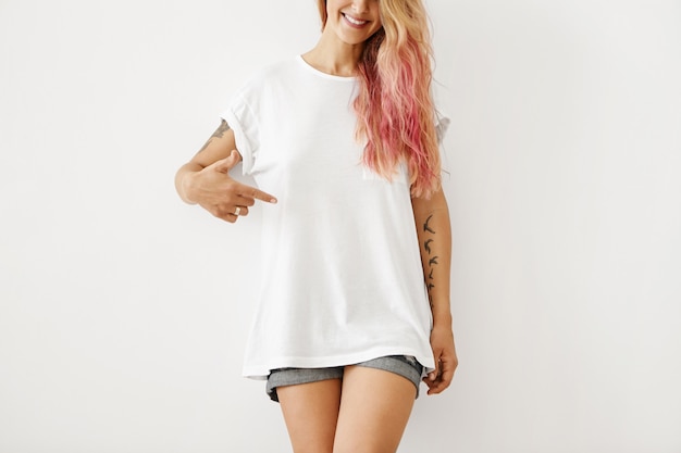 Free photo clothing, design, style, fashion and advertising. portrait of unrecognizable slim hipster woman with pink hair highlights and tattooed arms smiling, pointing finger at blank t-shirt