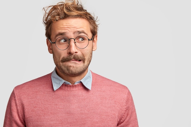 Free Photo clueless confused caucasian male bites lower lip and looks with anxious expression aside, worries before something important in life, make decision, dressed casually, isolated over white wall