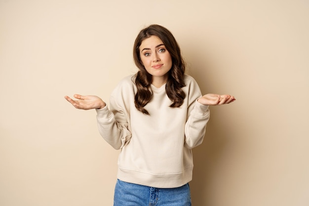 Clueless cute girl shrugging and smiling standing over beige background