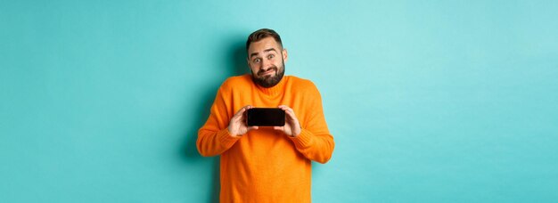 Clueless guy shrugging and showing mobile screen indecisive emotion standing in orange sweater over