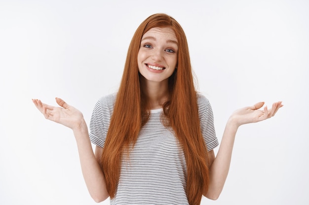 Clueless lovely tender redhead blue-eyed young girl smiling unaware shrugging hands spread sideways have no clue questioned apologize cannot answer, standing doubtful white wall