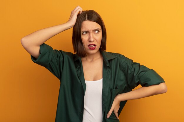 Clueless pretty woman puts hand on head and looks at side isolated on orange wall