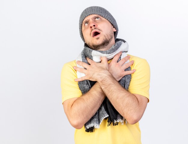 Clueless young blonde ill slavic man wearing winter hat and scarf puts hands on chest looking up isolated on white wall with copy space