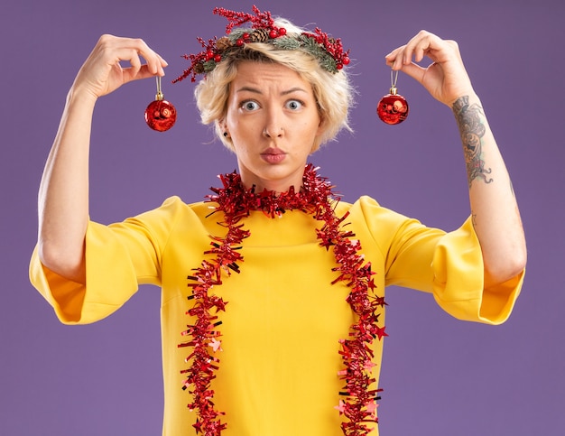 Free Photo clueless young blonde woman wearing christmas head wreath and tinsel garland around neck looking at camera holding christmas baubles near head isolated on purple background