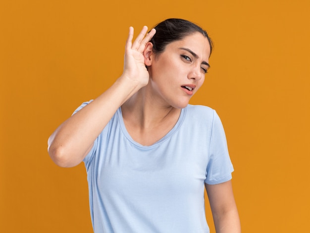 Clueless young brunette caucasian girl holds hand behind ear on orange